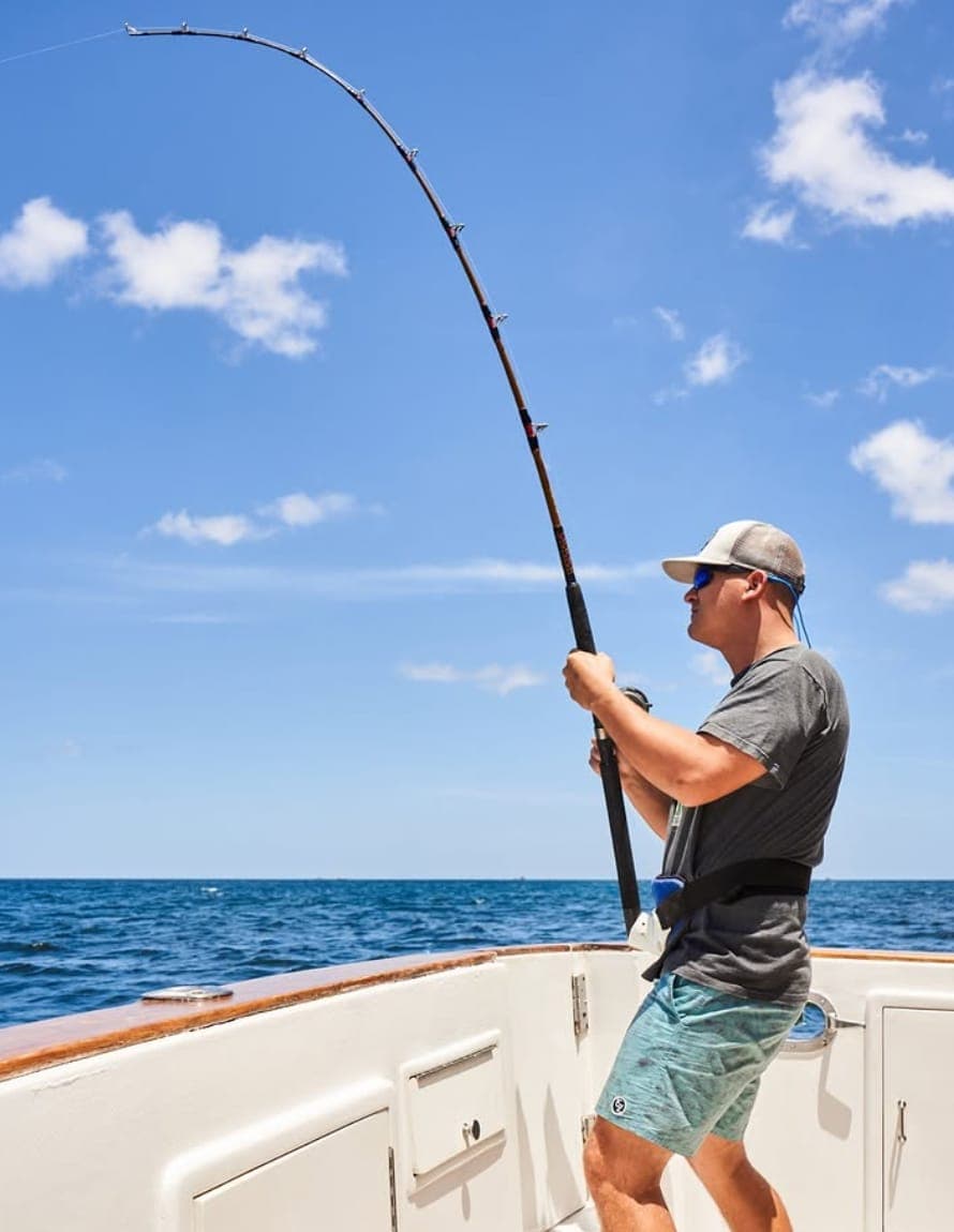 An angler leaning back on a bent rod, fighting a fish from the stern of the boat under a blue sky
