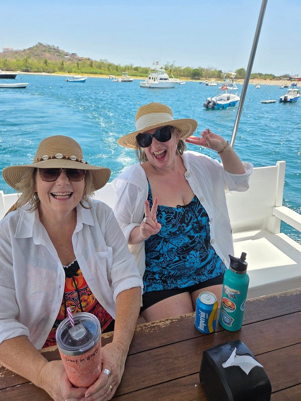 Two smiling guests in sun hats at a table on deck, with boats moored in the bay behind them
