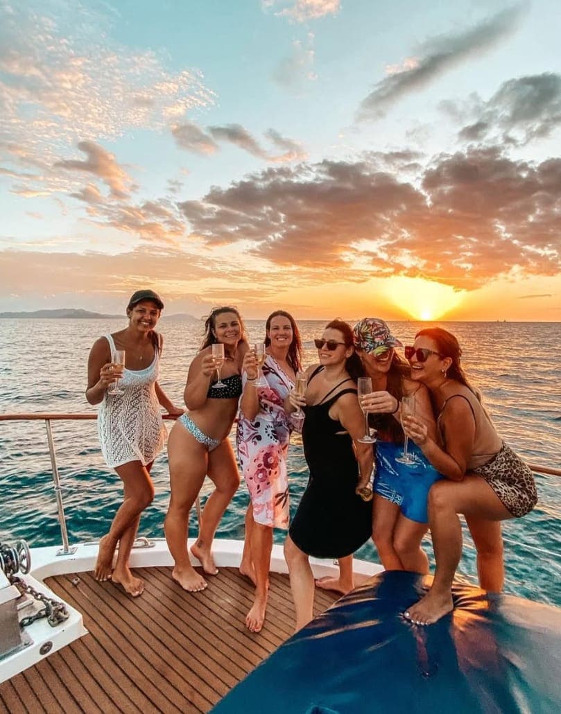 A group of friends raising glasses on the bow as the sun sets over the Pacific