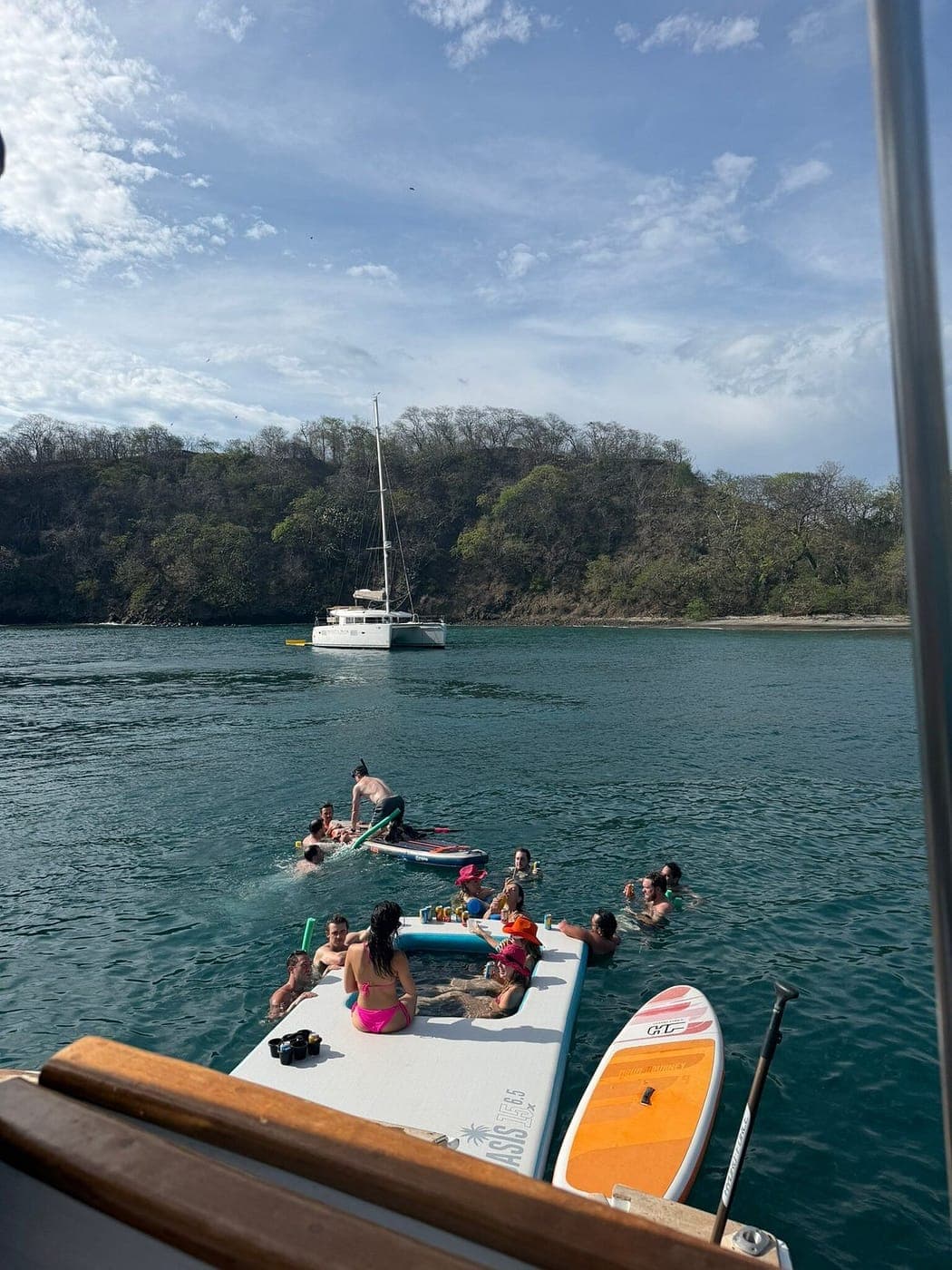 Guests swimming and lounging on a floating mat beside paddleboards in a quiet green cove