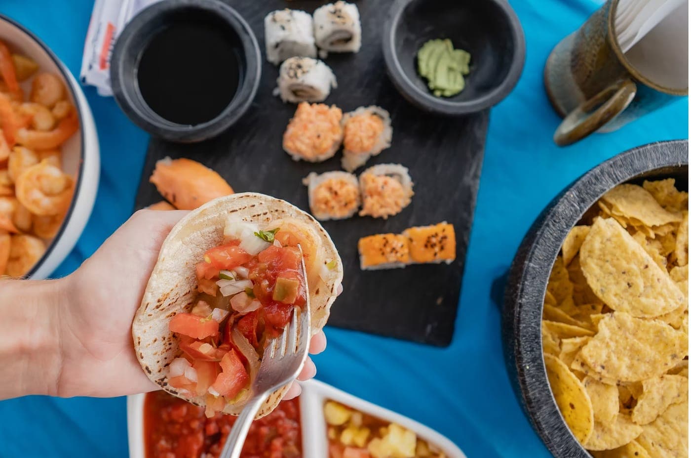 A hand holding a fresh taco over a table of sushi, shrimp and tortilla chips