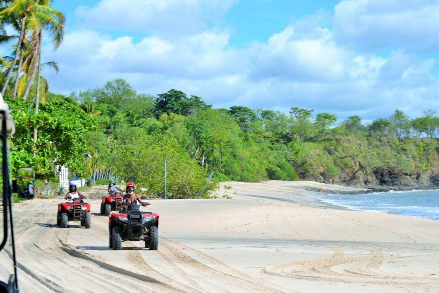 ATVs riding along a sandy beach road lined with palm trees