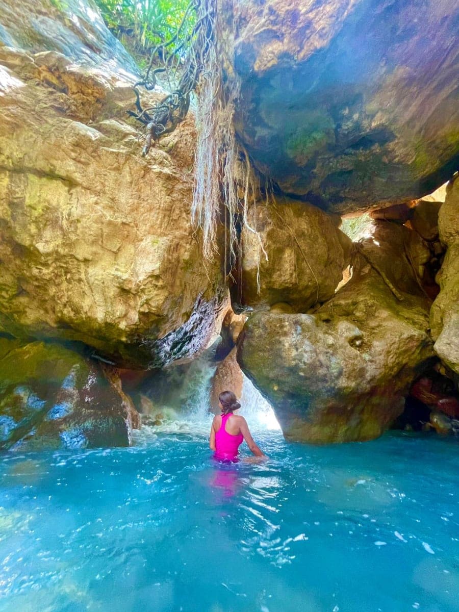 A swimmer in turquoise water inside a rock canyon beneath a thin waterfall
